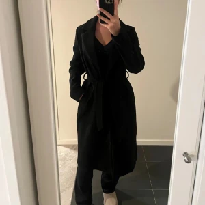 Svart kappa med bälte - Säljer en elegant svart kappa i mycket bra skick. Den har långa ärmar och ett bälte i midjan för en snygg passform. Perfekt för höst och vinter, både stilren och varm. Passar till alla tillfällen!
