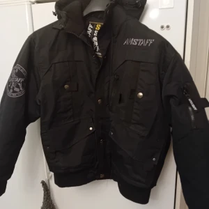 Svart jacka från Amstaff - Säljer en svart jacka från Amstaff i mycket bra skick. Jackan har en cool design med en stor hundlogga på ryggen och flera praktiska fickor framtill. Den är perfekt för höst och vinter med sin varma och robusta stil. Dragkedja och knappar framtill för extra skydd mot kyla.