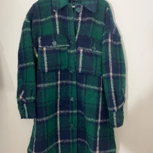 Grön och blå rutig kappa från Gina Tricot - Säljer en superfin grön och blå rutig kappa från Gina Tricot i storlek M. Den har långa ärmar och knappar framtill. Perfekt för höst och vår när du vill vara både varm och stilren. Två stora fickor på bröstet ger en cool look. Passar perfekt till både vardag och fest!