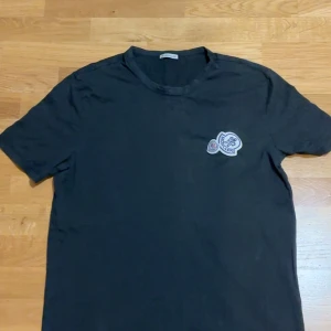 Svart t-shirt från Moncler - Säljer en snygg svart t-shirt från Moncler i storlek xl men passar M då den sitter lite mer slim. Passar bra om du är runt 175cm + den är väldigt bra skick. Den har en cool broderad logga på bröstet och är perfekt för både vardag och fest. Gjord i mjukt bomullsmaterial för extra komfort. Passar perfekt till jeans eller shorts!