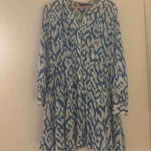 Blå och vit mönstrad klänning - Säljer en superfin blå och vit mönstrad klänning med långa ärmar. Den har en lös passform och är perfekt för både vår och sommar. Klänningen har ett unikt mönster som verkligen sticker ut. 