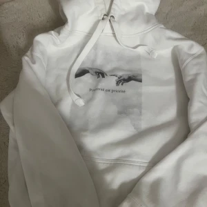 Vit hoodie med tryck - Säljer en vit hoodie med ett coolt tryck av två händer och texten 'Positivité est priorité'. Den är supermjuk och perfekt för chilliga dagar. Har en klassisk känguruficka och justerbar huva med snören. Passar perfekt till jeans eller mysbyxor!
