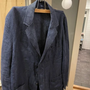 Blå linnekavaj från Grant Thomas - Säljer en stilren blå kavaj i 100% linne från Grant Thomas. Kavajen har en klassisk design med knappar framtill och fickor. Passar både till vardags och festliga tillfällen. Stor i storleken så snarare L än M. Mycket bra skick:)