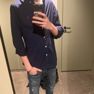 Ralph Lauren skjorta - Säljer en snygg mörkblå skjorta från Polo Ralph Lauren i storlek M. Skjortan är i nyskick och har en klassisk design med knappar och en broderad logga i rött på bröstet. Perfekt för både vardag och fest! 💙🍾