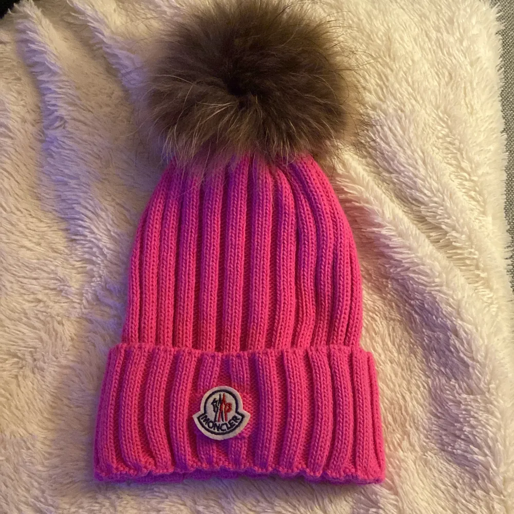 Superfin rosa stickad mössa från Moncler med en stor fluffig pälsboll på toppen. Den har en ribbad design och Monclers logga framtill. Perfekt för att hålla sig varm och snygg under kalla vinterdagar. 🧤❄️. Accessoarer.