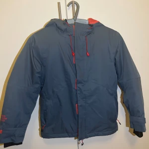 Blå och röd jacka från Neomondo - Säljer en snygg blå jacka från Neomondo med röda detaljer. Jackan har en huva och dragkedja framtill. Perfekt för kyliga dagar med sitt bekväma och funktionella design. Passar bra för både vardag och utomhusaktiviteter.