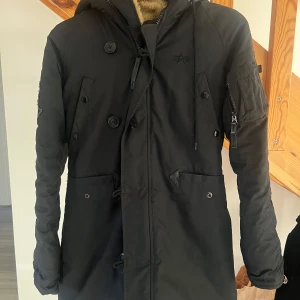 Svart parkajacka med foder - Säljer en svart Alpha Industires jacka med foder och huva (fejkpälsen finns). Jackan har långa ärmar och stängs med både dragkedja och knappar. Perfekt för kalla vinterdagar. Den har praktiska fickor och en stilren design❤️