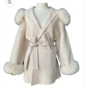 Beige kappa med pälsdetaljer - Superfin beige kappa med fluffiga pälsdetaljer på huva och ärmslut. Den har ett bälte i midjan och fickor fram. Änvänd ändas två gånger. Pris kan diskuteras.