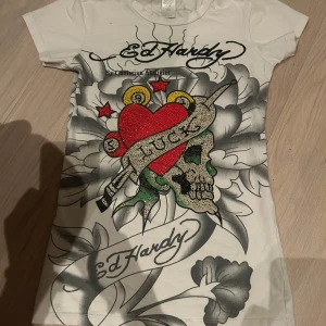 Vit Ed Hardy t-shirt med tryck - Säljer en cool vit t-shirt från Ed Hardy med ett stort, färgglatt tryck av ett hjärta, dödskalle och blommor. Texten 'Luck' är tryckt över hjärtat. Perfekt för dig som gillar y2k stil!