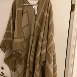 Beige poncho med fransar - Säljer en superfin beige poncho med ett geometriskt mönster i ljusare nyanser. Den har fransar längs nederkanten som ger en bohemisk touch. Perfekt för höst och vår när du vill ha något lätt men ändå värmande. Passar till både vardag och fest!