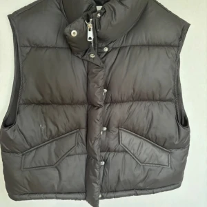 Svart dunväst med dragkedja - Säljer en svart dunväst i bra skick. Västen har en dragkedja och knappar framtill samt två praktiska fickor. Perfekt för höst och vinter när du vill hålla dig varm utan att bära en jacka. Passar både till vardags och utomhusaktiviteter.