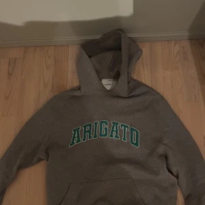 Grå hoodie med texten 'ARIGATO' - Säljer en grå hoodie från Axel Arigato med tryck. Den är superbekväm och perfekt för både chill och vardag. Hoodien har en klassisk passform med en stor ficka fram och en mysig huva. Perfekt för höst och vinter! Köptes ny för 1600. Snörrerna vid luvan saknas, annars bra skick.