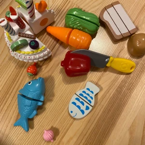Lekmat i plast - En samling färgglada leksaksmatvaror i plast, inklusive en tårta med ljus, en blå fisk, en morot, en paprika och en kniv. Perfekt för låtsaslek i köket! Maten kan delas då den sitter ihop med kardborre. Tyvärr saknas vissa delar, men det är lekbart ändå! 