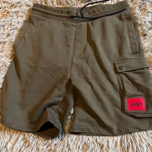 Olivgröna shorts från Hugo - Säljer ett par olivgröna shorts från Hugo i nyskick. De har en bekväm resårmidja med dragsko och en praktisk ficka med knapp på sidan. Perfekta för en avslappnad stil i sommar! 🟢