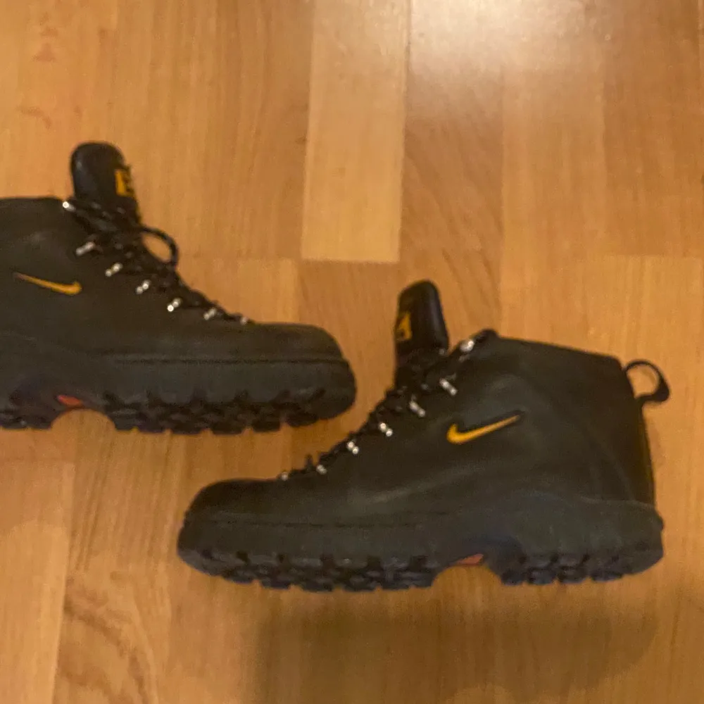 Säljer ett par svarta Nike boots i nyskick. De har en robust design med snörning och en liten gul Nike-logga på sidan. Perfekta för höst och vinter med sin slitstarka sula och bekväma passform. Skorna är både snygga och funktionella för vardagsbruk.. Skor.