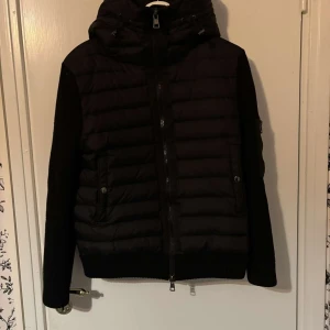 Svart dunjacka från Moncler - Säljer en snygg svart dunjacka från Moncler. Jackan har en quiltad design med dragkedja och en huva för extra värme. Perfekt för kyliga höst- och vinterdagar. Den har långa ärmar och en stilren look. Passar både till vardags och lite finare tillfällen.