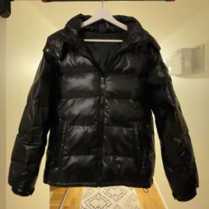 Svart pufferjacka - Säljer en svart pufferjacka i mycket bra /Ny skick. Har använt den bara 1 gång. Den är perfekt för vintern med sin tjocka vaddering och har en praktisk dragkedja framtill. Jackan har långa ärmar och en huva för extra värme. Den är både snygg och funktionell, perfekt för kalla dagar!