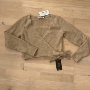 Beige omlottkofta från NA-KD - Säljer en superfin beige omlottkofta från NA-KD. Den är croppad och har en snygg knytning i sidan. Perfekt för höst och vår. Koftan är i storlek XS och är helt nytt skick då den aldrig är använd.🧡