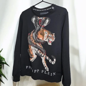 Svart tröja med tiger- och ormtryck från Philipp Plein - Säljer en svart tröja från Philipp Plein med ett coolt tryck av en tiger och orm på framsidan. Tröjan har långa ärmar och är perfekt för höst och vinter. Den är i bra skick och har en snygg passform. Perfekt för dig som vill sticka ut!