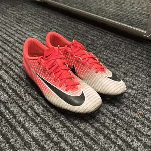 Nike Mercurial fotbollsskor - Säljer ett par Nike Mercurial fotbollsskor i rött och vitt med svarta detaljer. Skorna har snörning och är i bra skick, perfekta för fotbollsträning eller match. De har en snygg design med Nike-loggan på sidan och dobbar för bra grepp på planen.