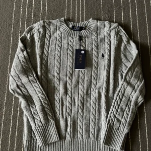 Grå stickad tröja från Polo Ralph Lauren - Säljer en snygg grå stickad tröja från Polo Ralph Lauren. Den har ett klassiskt flätmönster och är i helt nytt skick med etikett kvar. Perfekt för kyliga dagar och passar både till vardags och lite finare tillfällen.