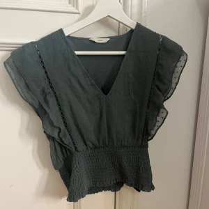 Grön v-ringad topp från ONLY - Säljer en superfin grön topp från NA-KD. Den har en v-ringning och är ärmlös med volangdetaljer. Toppen är croppad och har en smockad nederdel som ger en snygg passform. Perfekt för både vardag och fest! 💚