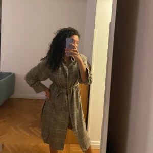 Rutig klänning med bälte - Säljer en superfin rutig jacka med bälte i midjan. Den har långa ärmar och en klassisk krage. Perfekt för höst och vår, och kan stylas både upp och ner beroende på tillfälle. Jackan är i en neutral färgton som passar till det mesta. 🖤