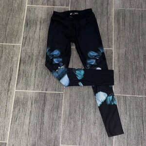 Svarta leggings med blått mönster från SOC - Snygga svarta leggings från SOC med ett coolt blått mönster. De har en tight passform och är gjorda i ett syntetmaterial som andas. Perfekta för både gymmet och löprundan!