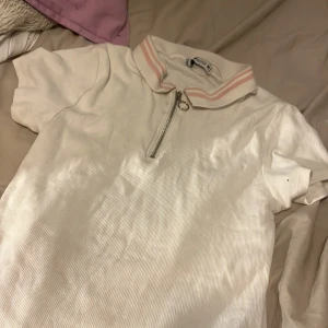 Vit ribbad polotopp från Sister - Säljer en vit ribbad polotopp från new yorker i storlek M. Toppen har en dragkedja framtill och korta ärmar. Kragen har en söt rosa randig detalj. Perfekt för en avslappnad stil eller vardagsbruk.