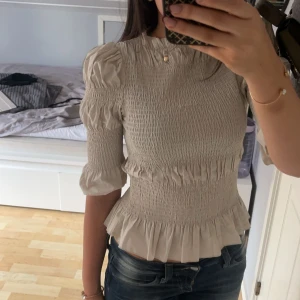 Beige blus med volangdetaljer - Säljer en superfin beige blus med volangdetaljer. Den har trekvartsärmar och en smockad design som ger en tight passform. Perfekt för både vardag och fest! 🥰säljer pga inte kommer till användning men tycker den är skitcool💕
