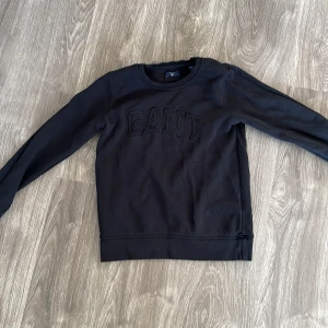 Svart sweatshirt från GANT - Säljer en stilren svart sweatshirt från GANT i storlek XS. Tröjan har en diskret logga på bröstet och är perfekt för både vardag och avslappnade tillfällen. Den är i mycket bra skick och gjord av mjukt material för extra komfort.