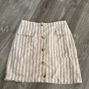 Beige och vit randig kjol med knappar - Säljer en superfin beige och vit randig kjol med stora knappar framtill. Kjolen har praktiska fickor och är perfekt för sommardagar. Den är i ett lätt och bekvämt material, perfekt för en avslappnad stil. Passar bra till både vardag och fest!