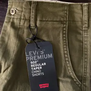 Säljer ett par snygga olivgröna Levi's Premium 502 Regular Taper Chino Shorts. De är i nytt skick och har en klassisk design med knappar och fickor. Perfekta för sommaren och vårens alla äventyr. Passar både till vardags och lite mer uppklädda tillfällen. Storlek 28.