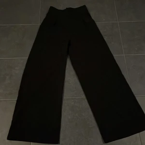 Svarta kostymbyxor från NA-KD - Säljer ett par stilrena svarta kostymbyxor från NA-KD. De är högmidjade och har en loose fit med vida ben, perfekt för både vardag och fest. Materialet känns mjukt och bekvämt, vilket gör dem sköna att bära hela dagen. Passar bra till både sneakers och klackar!