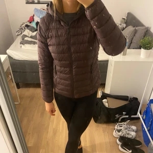 Brun/lila dunjacka med huva - Säljer en snygg och varm dunjacka med huva. Perfekt för kyliga höst- och vinterdagar. Jackan har en dragkedja framtill och är långärmad. Den är i bra skick och passar perfekt för både vardag och utomhusaktiviteter.