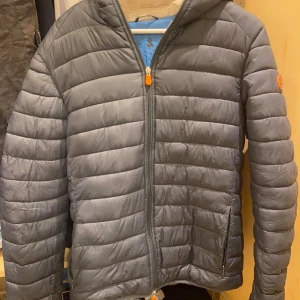 Save the duck jacka - Säljer en snygg grå pufferjacka med huva. Jackan har en dragkedja framtill och är perfekt för kyliga dagar. Den är fodrad med mjukt material för extra värme och komfort. Perfekt för höst och vinter. Pris kan diskuteras vid en snabb affär!