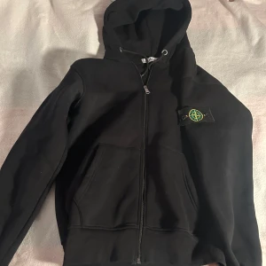 Svart hoodie från Stone Island - Säljer en svart hoodie från Stone Island i bra skick. Den har en dragkedja framtill och en klassisk logga på ärmen. Perfekt för höst och vinter, och den är superbekväm med sin mjuka insida. Passar både till vardags och chillkvällar. Hör av dig vid intresse!