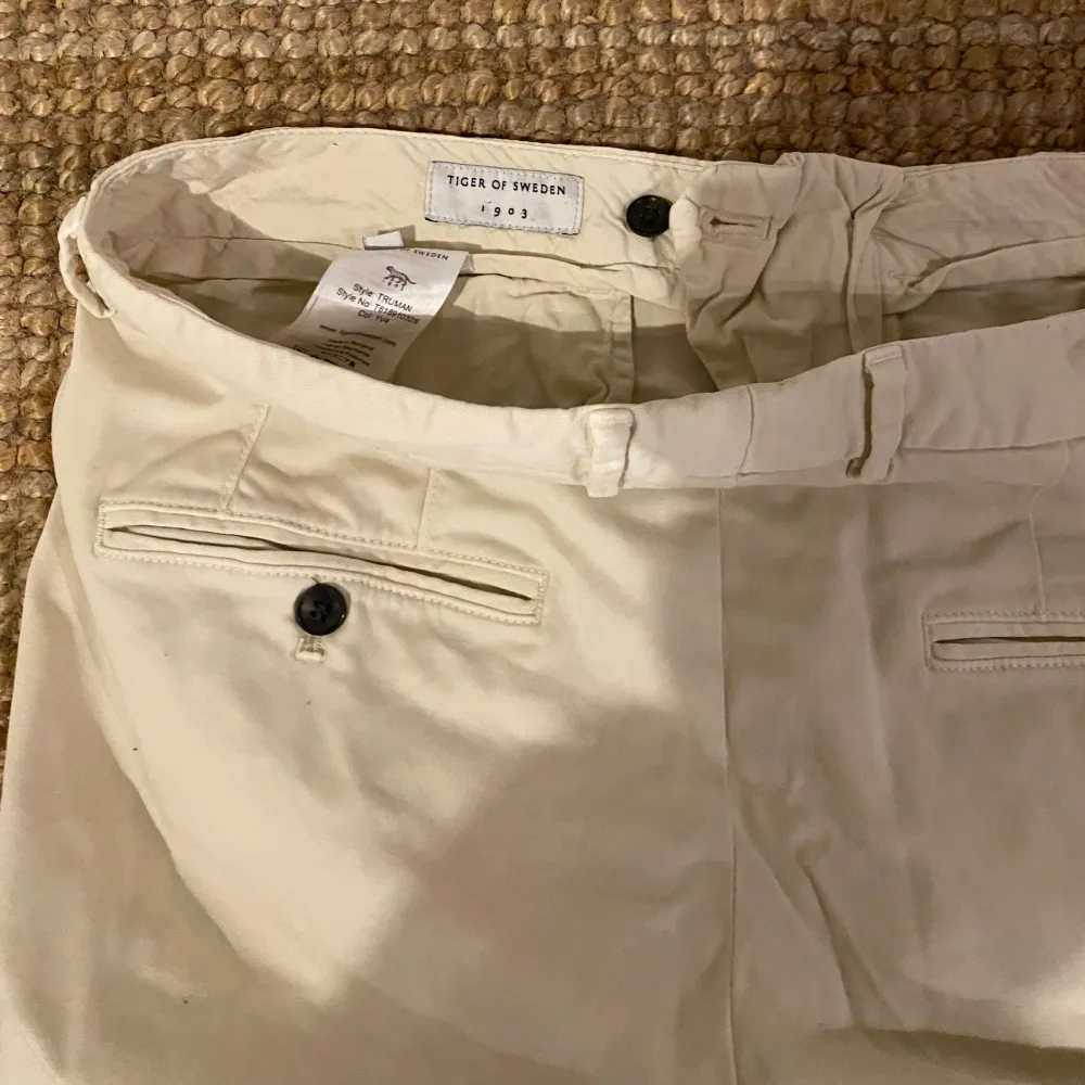 Säljer ett par stilrena beige chinos från Tiger of Sweden i storlek 52. De är i mycket bra skick och perfekta för både vardag och fest. Byxorna har en normal passform med knappar och dragkedja i midjan. De är tillverkade i ett mjukt bomullsmaterial som gör dem bekväma att bära hela dagen.. Jeans & Byxor.