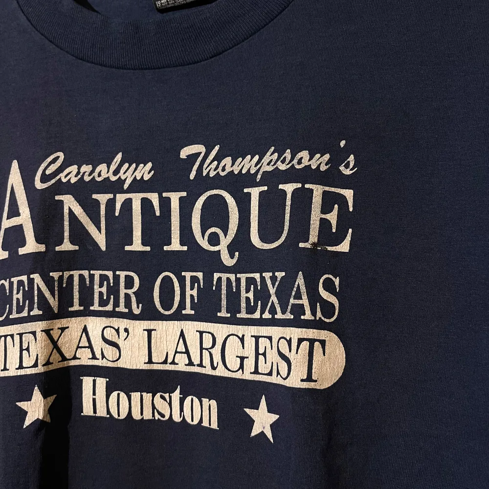 Säljer en vintage  mörkblå t-shirt från Fruit of the Loom med tryck 'Antique Center of Texas' i vitt. Den är i bra skick och gjord av 50% bomull och 50% polyester. Perfekt för en avslappnad stil eller som souvenir från Houston!. T-shirts.