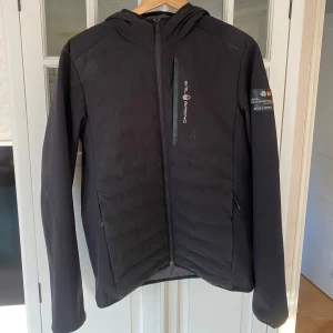 Svart jacka från Sail Racing - Säljer en svart jacka från Sail Racing i mycket bra skick. Den har en stilren design med dragkedja och huva, perfekt för höst och vår. Jackan är långärmad och har en logga på ärmen och huvan. Passar både till vardags och lite mer sportiga aktiviteter.