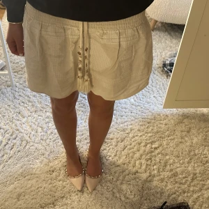 Zara kjol - Säljer en superfin vit kortkjol med knytband framtill. Kjolen har en avslappnad passform och är perfekt för sommardagar. Den är gjord i ett lätt och bekvämt material, vilket gör den idealisk för både vardag och fest. Passar perfekt med en enkel topp och sandaler!