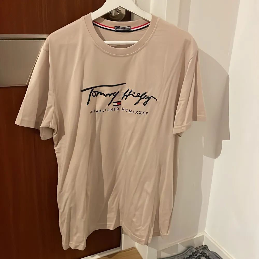 Säljer en snygg beige t-shirt från Tommy Hilfiger i mycket bra skick. Den har ett stilrent tryck på framsidan med märkets logga och text. Perfekt för en avslappnad stil och passar till både vardag och fest. Storleken är normal och materialet känns mjukt och skönt mot huden.. T-shirts.