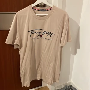 Beige t-shirt från Tommy Hilfiger - Säljer en snygg beige t-shirt från Tommy Hilfiger i mycket bra skick. Den har ett stilrent tryck på framsidan med märkets logga och text. Perfekt för en avslappnad stil och passar till både vardag och fest. Storleken är normal och materialet känns mjukt och skönt mot huden.