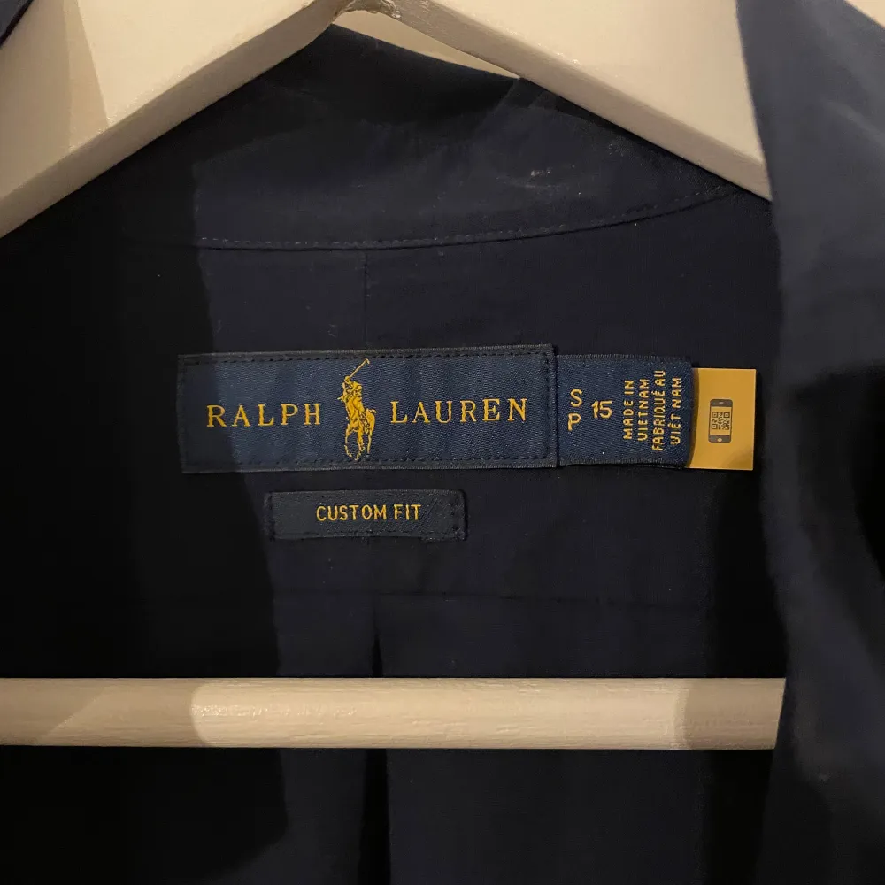 Säljer en stilren mörkblå skjorta från Ralph Lauren i custom fit. Skjortan har långa ärmar och klassiska knappar framtill. Perfekt för både vardag och festliga tillfällen. Passar bra till både jeans och kostymbyxor. Skjortan är i mycket bra skick och redo för nya äventyr!. Skjortor.