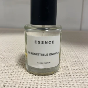 Essnce Irresistible Enigma Eau de Parfum - Säljer en flaska Essnce Irresistible Enigma Eau de Parfum. Flaskan är genomskinlig med en enkel vit etikett och svart text. De mängd på bild 2
