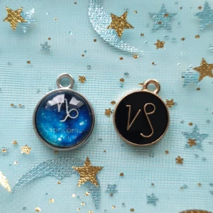 Zodiakberlocker med Stenbockens tecken - Två snygga berlocker med Stenbockens stjärntecken. Den ena har en blå bakgrund med stjärnhimmel och texten 'Capricorn', medan den andra har en svart bakgrund med en stilren symbol i guld. Perfekta för att göra ditt smycke personligt!