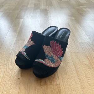 Svarta klackskor med broderade fåglar - Säljer ett par snygga svarta klackskor. De har en cool broderad design med färgglada fåglar i rött och blått på ovansidan. Skorna har en öppen tå och en stabil klack som ger bra höjd. Perfekta för en utekväll eller fest!
