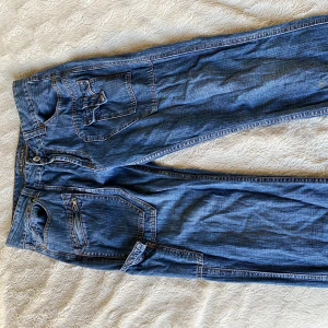 Blå jeans från Cailina - Säljer ett par snygga blå jeans från Cailina. De har en cool 80/90-tals design med flera fickor och detaljer som ger en unik look. Perfekta för vardags. Jeansen är i bra skick, innerbenet ungefär 80cm och lite över 40 över midjan (sista bild) 