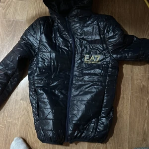 Mörkblå  quiltad jacka från armani  - Säljer en mörkblå quiltad jacka från Armani . Den är perfekt för kyliga dagar med sin pufferstil och har en praktisk huva. Jackan har en dragkedja framtill och är långärmad. Den är i bra skick och redo för nya äventyr!