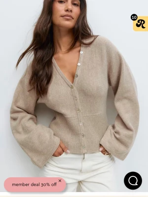 Beige kofta med pärlknappar - Slutsåld beige kofta från Gina med djup v-ringning och pärlknappar framtill. Koftan har breda, långa ärmar och ribbad midja som ger en snygg passform. Perfekt att styla med jeans eller kjol för en cool look. Tröjan är lite sönder i midjan men går att fixa enkelt💞